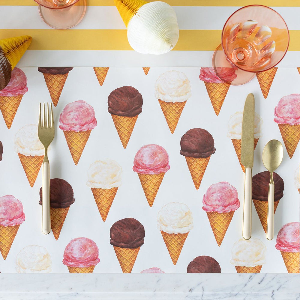 Placemat with ice cream cone pattern, cutlery, and pink glasses on a yellow and white striped background.