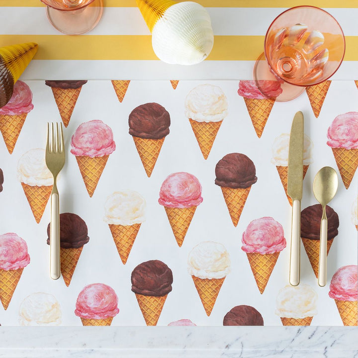 Placemat with ice cream cone pattern, cutlery, and pink glasses on a yellow and white striped background.