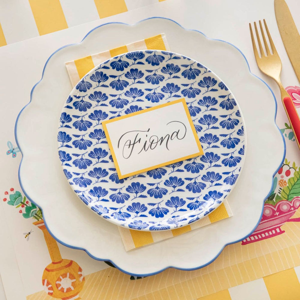 Decorative table setting with a blue floral plate, name card, and colorful cutlery on a yellow and white checkered tablecloth.