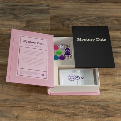 Open mystery date box with pink card and black lid on wooden surface