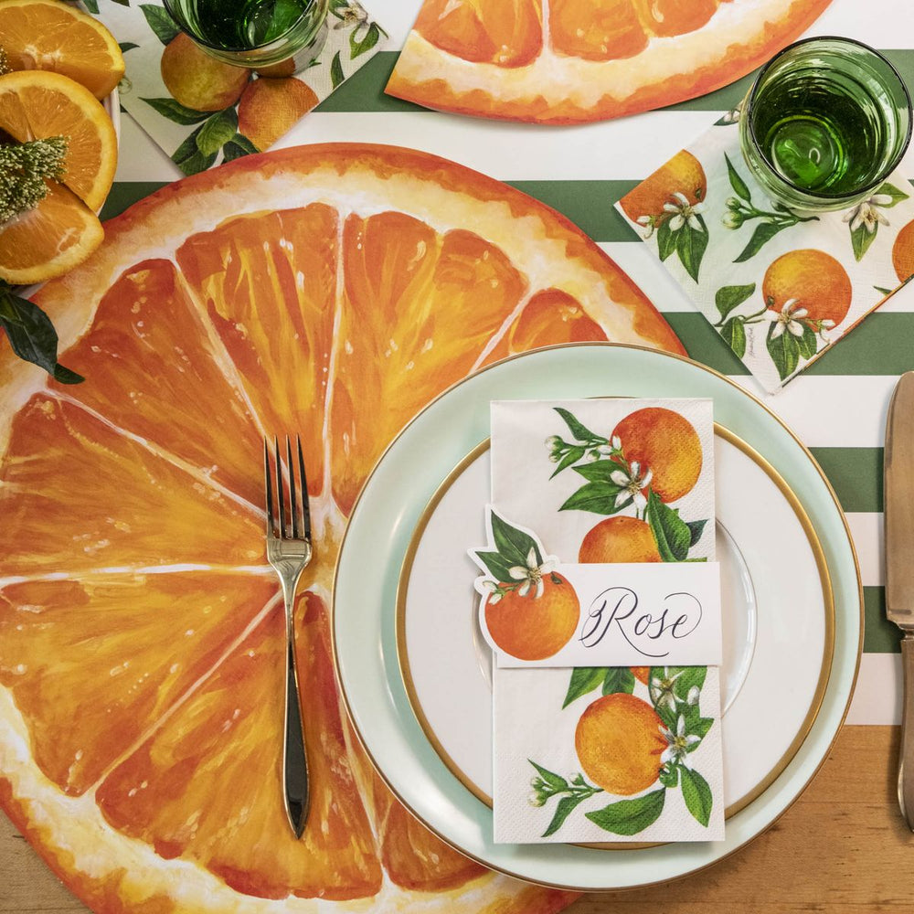 Table setting with orange-themed decor, including a large orange slice, a place card, and cutlery on a striped tablecloth.