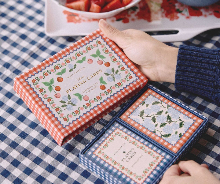 Hand holding a deck of playing cards with decorative packaging on a checkered tablecloth.