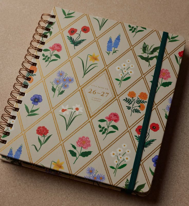 A spiral-bound planner or notebook with a cream cover decorated with a gold diamond grid and colorful floral illustrations. The cover includes a central label reading '26-27 JUNE - 27 JUNE' and features a dark green elastic closure band.