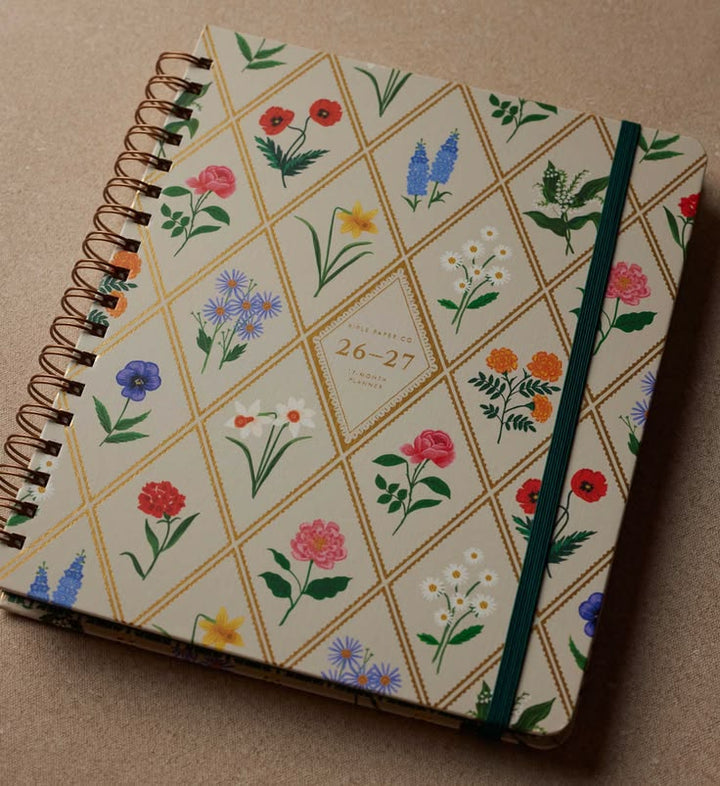 A spiral-bound planner or notebook with a cream cover decorated with a gold diamond grid and colorful floral illustrations. The cover includes a central label reading '26-27 JUNE - 27 JUNE' and features a dark green elastic closure band.