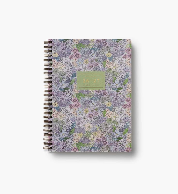 A spiral-bound 2027 daily planner with a dense floral pattern in shades of purple, lavender, and green, featuring a central sage green label with gold text.