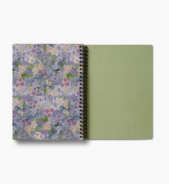 A spiral-bound notebook shown open on a white background, with a floral-patterned cover in lavender, purple, and green tones on the left and a solid sage green blank page on the right.