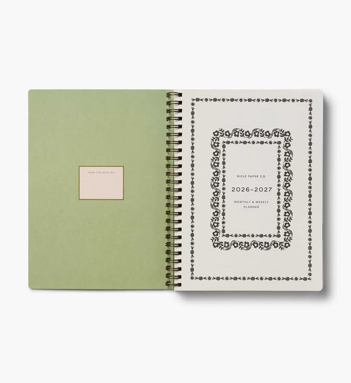 An open spiral-bound planner or notebook with a sage green cover on the left and an interior page with a black decorative border and text on the right, shown on a light gray background.