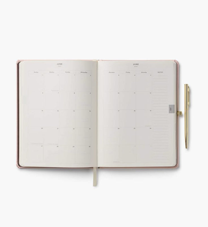 An open monthly planner with cream-colored pages and a slim gold pen resting on the right side, set against a clean white background.