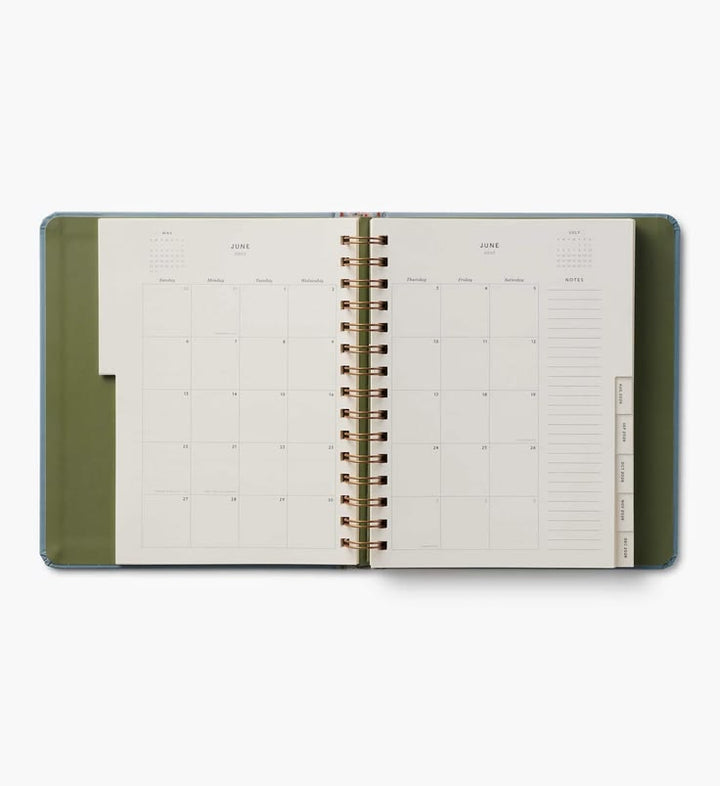 An open spiral-bound planner or calendar showing June and July monthly layouts, with an olive green cover and gold-toned spiral binding on a light gray background.