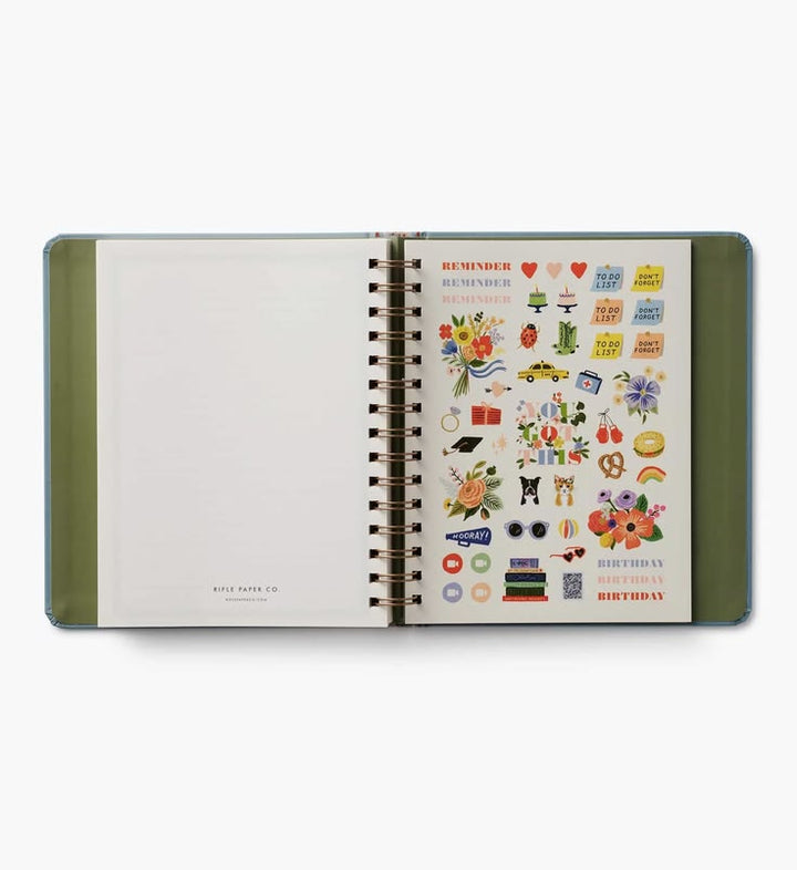 An open spiral-bound planner or notebook with an olive green cover, showing a decorated sticker sheet on the right page and blank pages on the left, photographed on a light gray background.