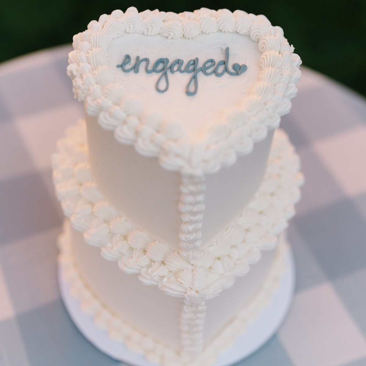 Two-tiered heart-shaped cake with 'engaged' decoration on a checkered tablecloth.
