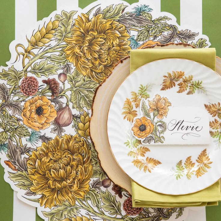 A styled table setting featuring a large white paper placemat with a detailed autumn floral illustration, layered under a round beige charger plate, a white paper place card reading 'Hirzo', a folded olive green napkin, and a matching smaller floral paper plate.
