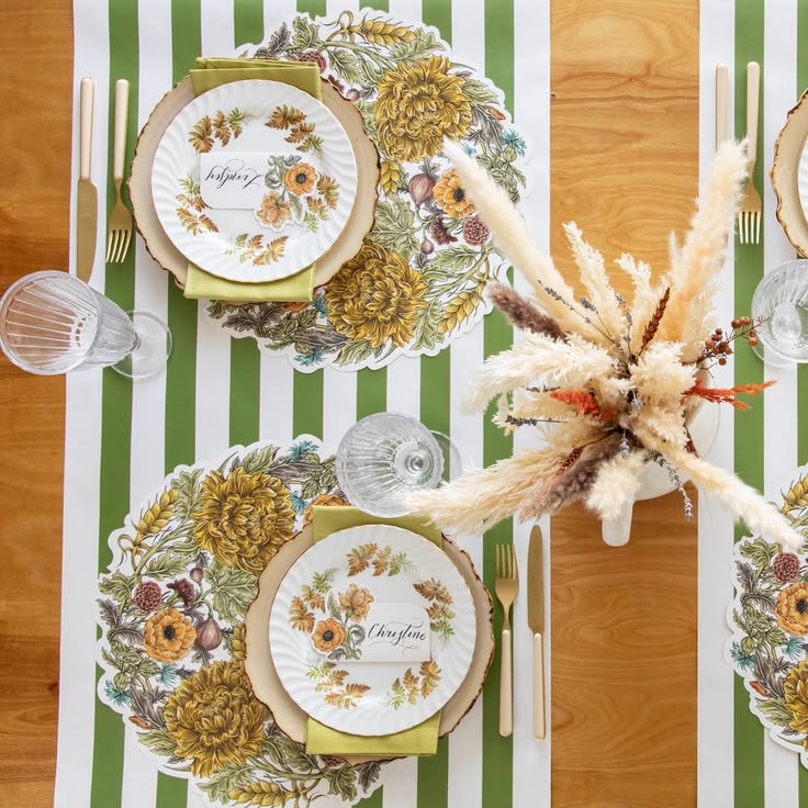 An overhead styled table setting featuring round floral placemats with a scalloped edge, white plates with autumn botanical illustrations, green napkins, clear glassware, and a centerpiece of dried pampas grass on a striped green, white, and wood table runner.
