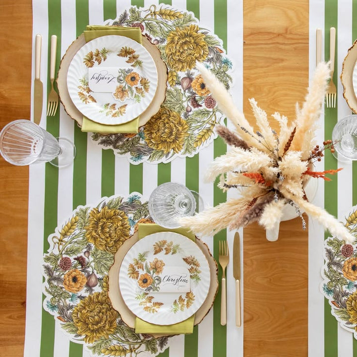 An overhead styled table setting featuring round floral placemats with a scalloped edge, white plates with autumn botanical illustrations, green napkins, clear glassware, and a centerpiece of dried pampas grass on a striped green, white, and wood table runner.