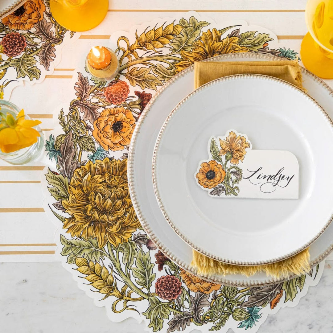 A styled table setting featuring a scalloped floral placemat with large yellow and orange flowers, green leaves, and gold accents, layered under stacked white plates with gold trim and a gold napkin. A small place card reading 'Lindsey' is centered on the top plate.