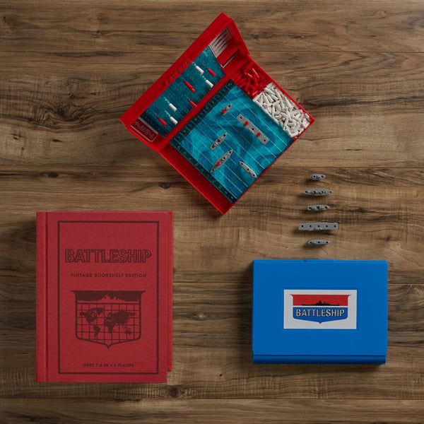 Battleship game components including a red box, blue tray, and cards on a wooden surface.