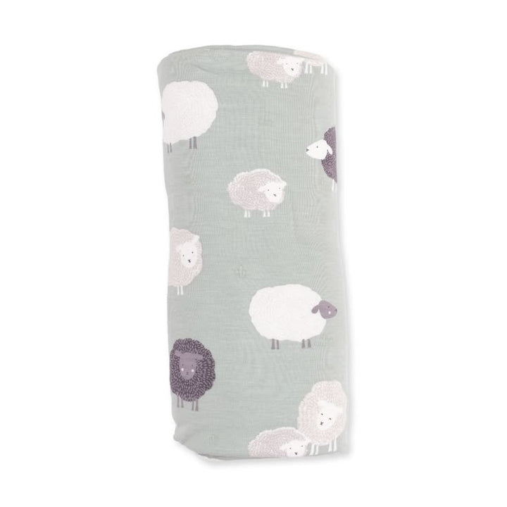 A rolled-up muslin blanket in a muted sage green color, featuring a pattern of stylized sheep in white, light pink, and dark purple, shown against a plain white background.
