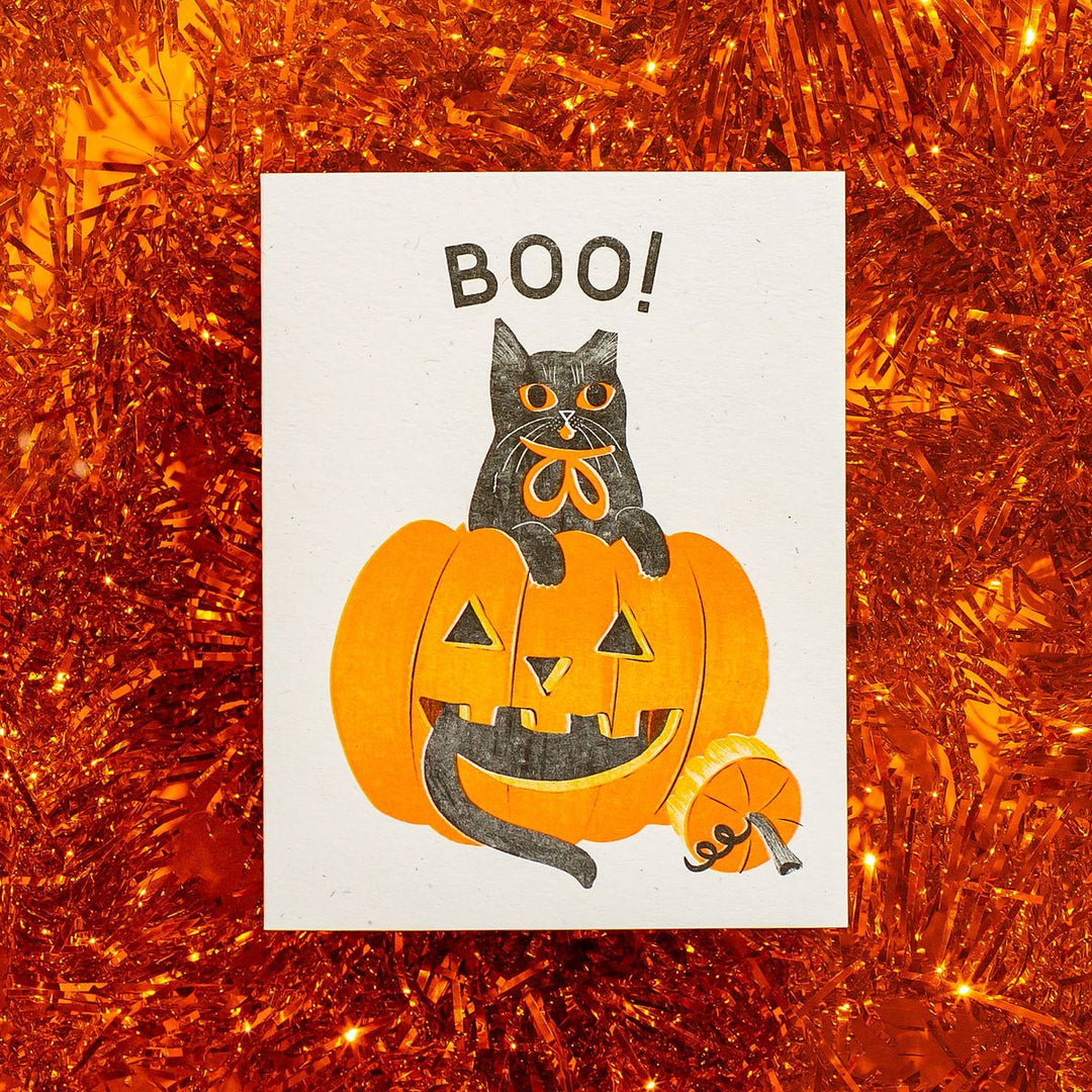 Halloween-themed card with a black cat on a pumpkin and 'BOO!' text, against an orange tinsel background.