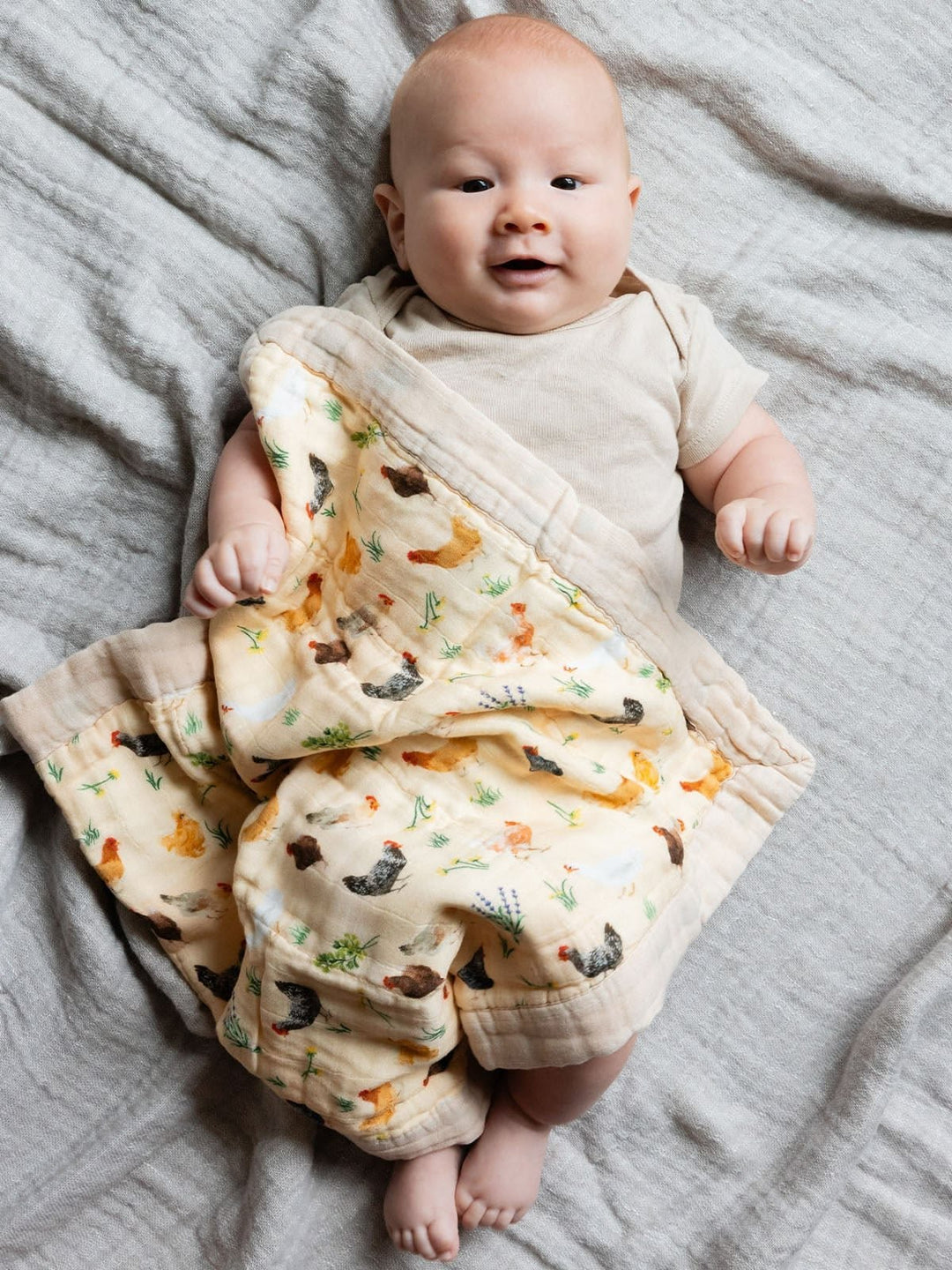 A baby lying on a textured gray blanket, wrapped in a cream-colored muslin swaddle blanket with a colorful forest and animal print.