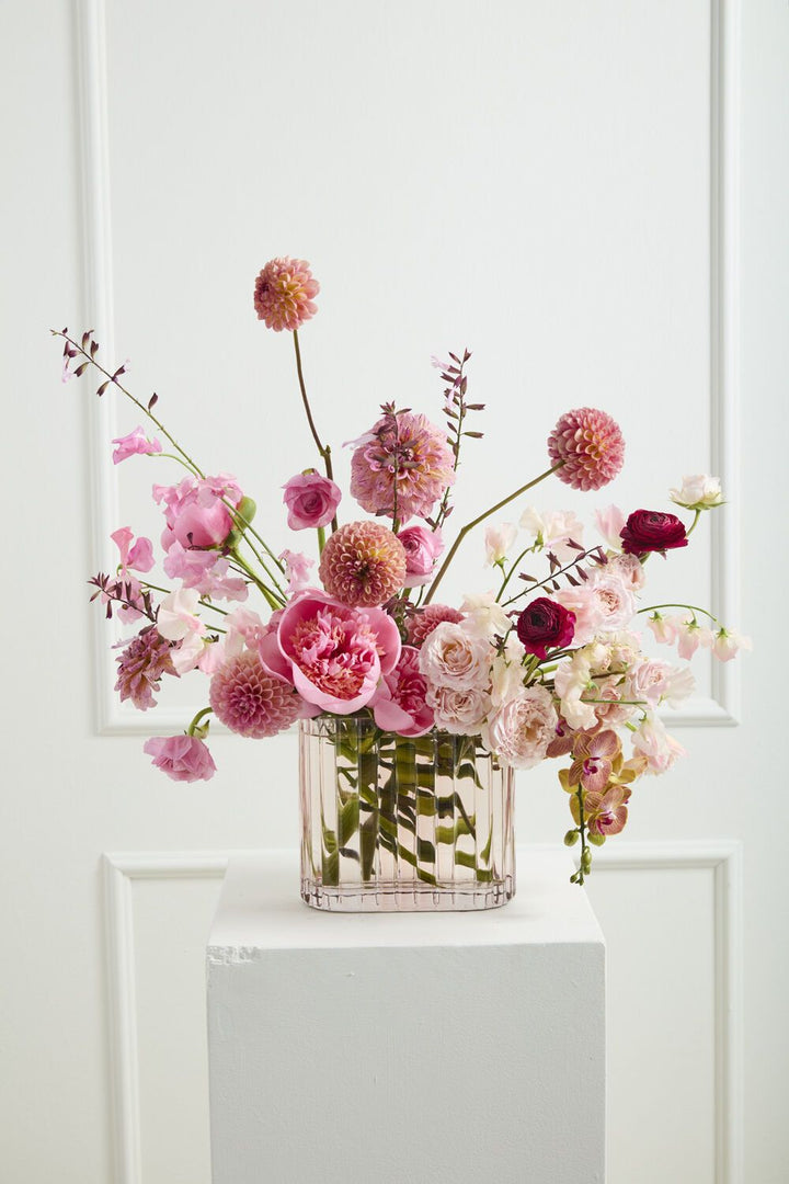 Bouquet of flowers in a clear vase on a white pedestal against a white paneled wall.
