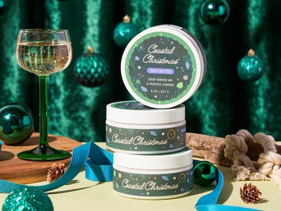 Three tins of 'Coastal Christmas' scented candles stacked on a table with a festive background.