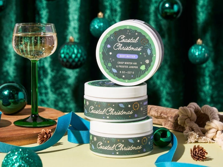 Three tins of 'Coastal Christmas' scented candles stacked on a table with a festive background.