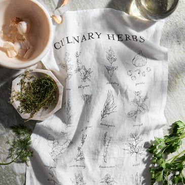 Culinary herbs towel with botanical illustrations on a table with fresh herbs and a cup.