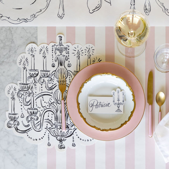Dinner table setting with pink plates, gold cutlery, and a decorative card on a striped tablecloth.