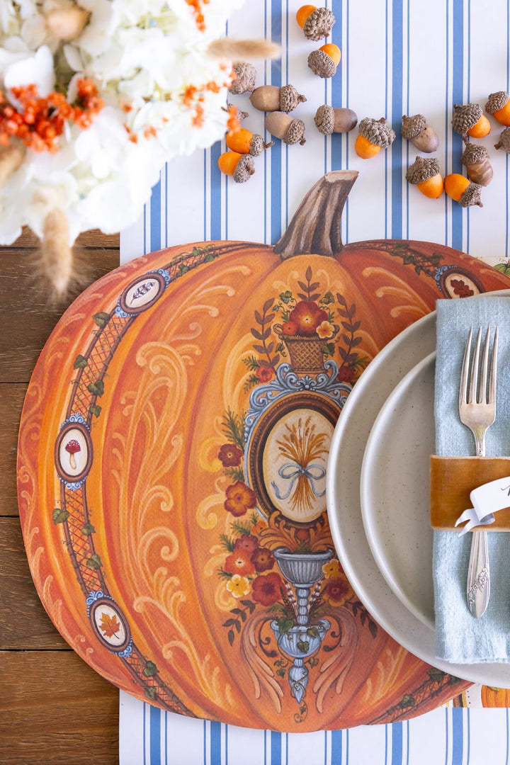 An autumn-themed round placemat featuring a detailed, colorful illustration of a decorated pumpkin, styled on a blue and white striped table runner with acorns, a white floral arrangement, and a place setting with a blue napkin and silver fork.