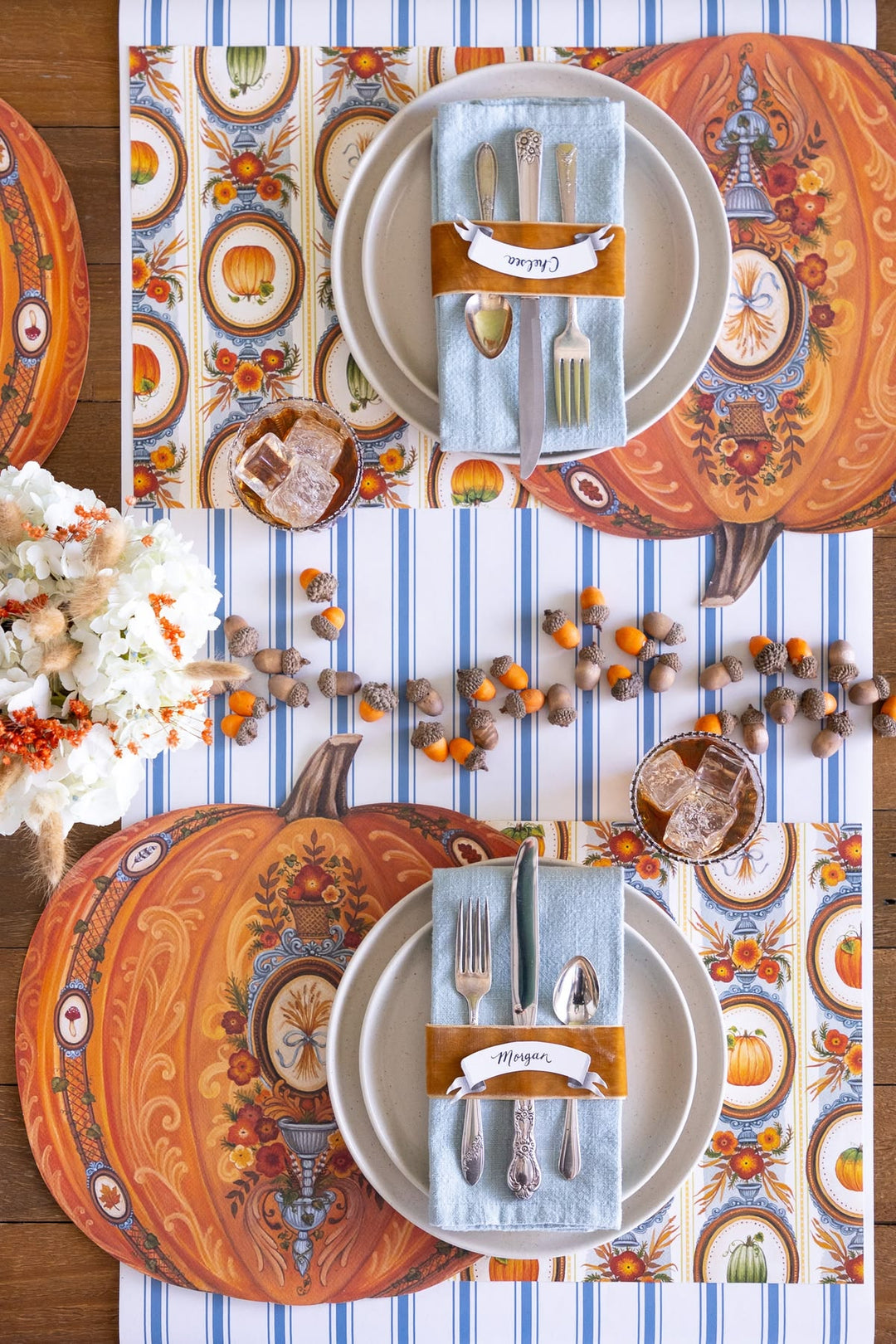 A styled autumn table setting with two place settings on a striped table runner. The setting includes orange patterned placemats with pumpkins, white plates, light blue napkins with silver cutlery, and orange fabric band wraps.