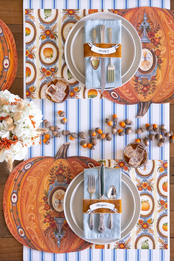 A styled autumn table setting with two place settings on a striped table runner. The setting includes orange patterned placemats with pumpkins, white plates, light blue napkins with silver cutlery, and orange fabric band wraps.