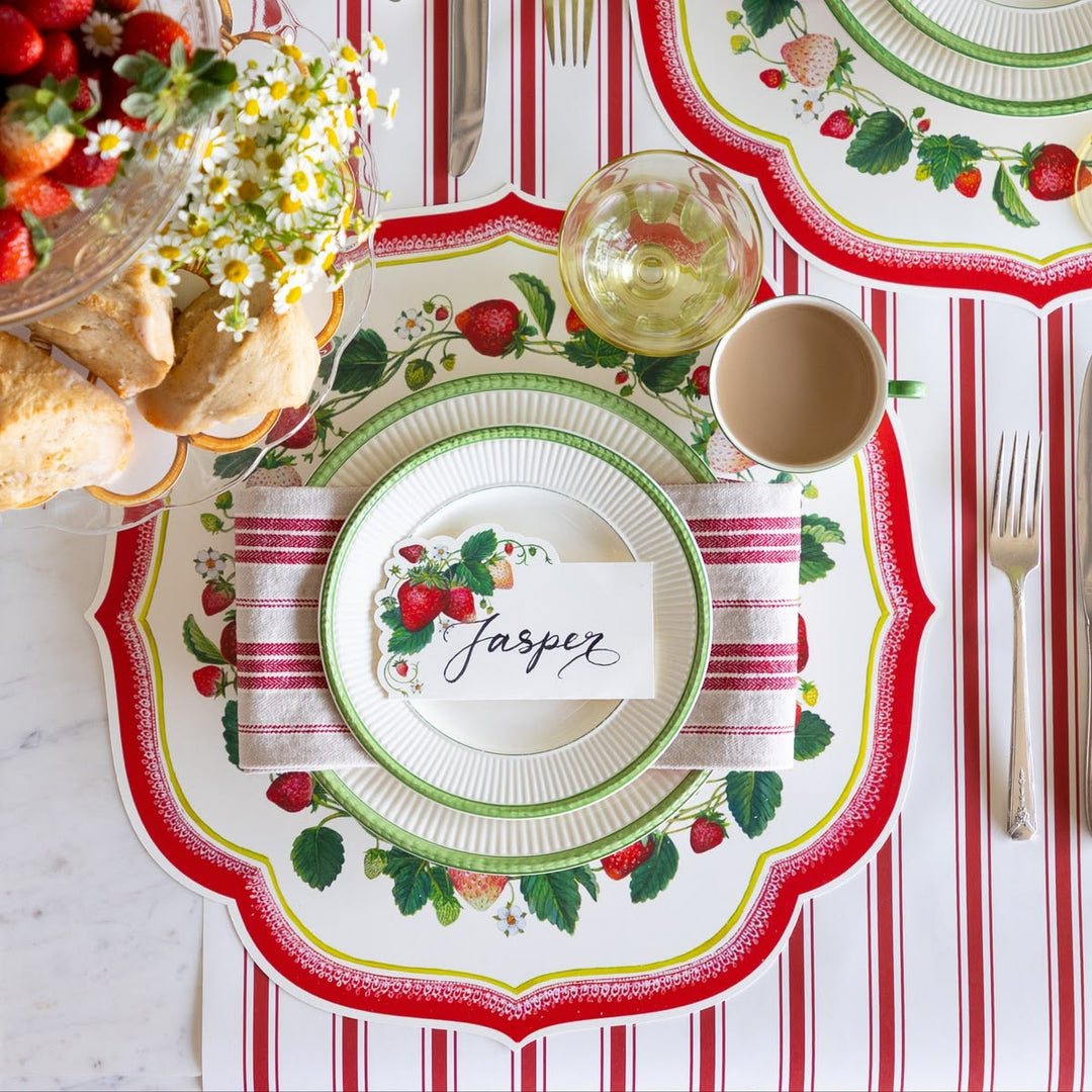 Decorative table setting with strawberry-themed plates and place card on a red and white striped tablecloth.