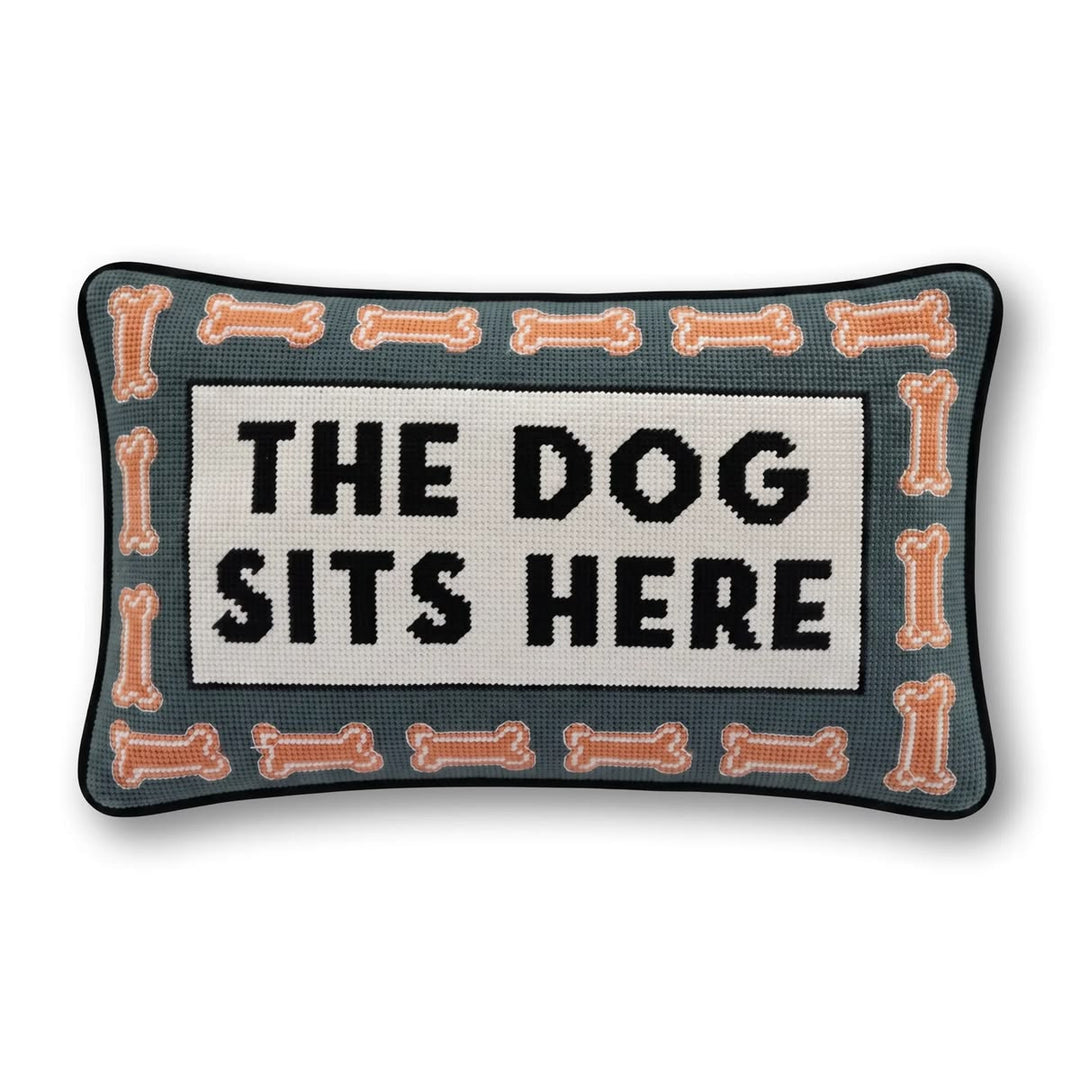 Rectangular decorative needlepoint pillow with the text 'THE DOG SITS HERE' in black on a cream center panel, surrounded by a border of peach-colored bones on a dark teal background.
