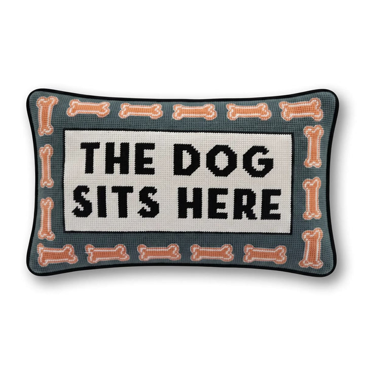 Rectangular decorative needlepoint pillow with the text 'THE DOG SITS HERE' in black on a cream center panel, surrounded by a border of peach-colored bones on a dark teal background.