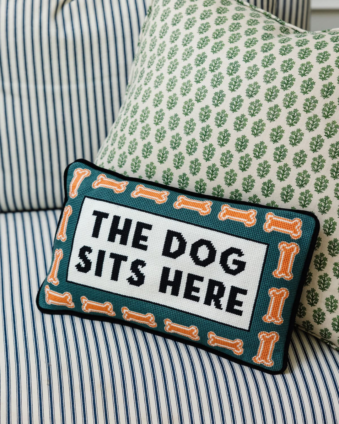 A rectangular embroidered throw pillow with the text 'THE DOG SITS HERE' in black on a white center panel, surrounded by an orange bone border and dark teal background. It is styled on a striped blue and white sofa next to a green patterned pillow.