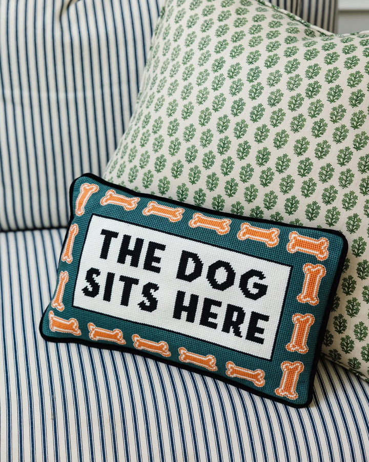 A rectangular embroidered throw pillow with the text 'THE DOG SITS HERE' in black on a white center panel, surrounded by an orange bone border and dark teal background. It is styled on a striped blue and white sofa next to a green patterned pillow.