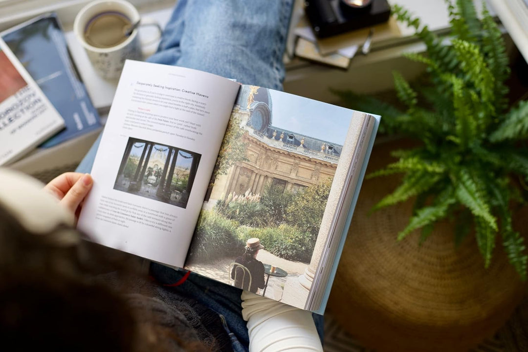 Person reading a book with a scenic image on a table with a plant and coffee cup.