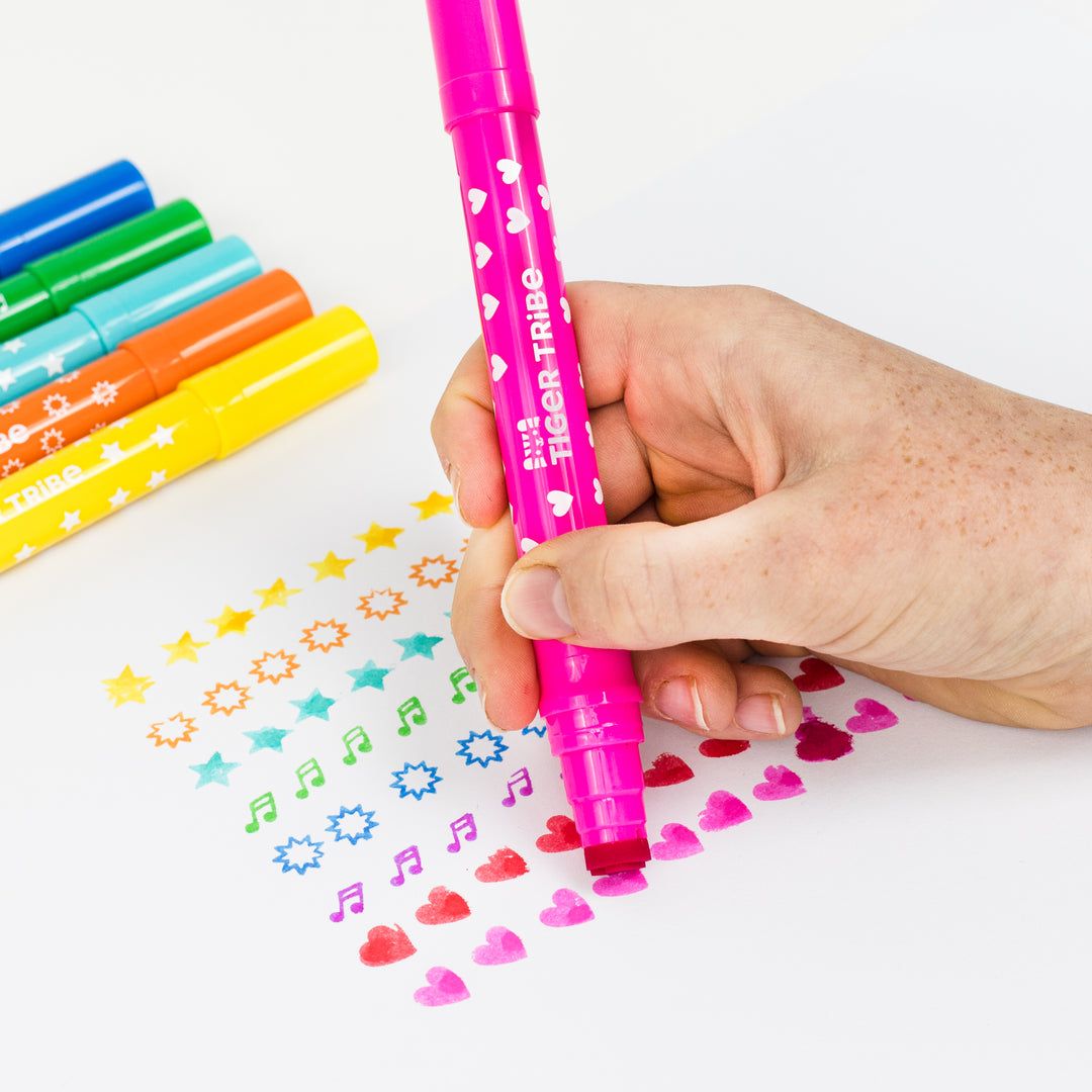 Hand holding a pink marker with colorful markers and stamped designs on a white background