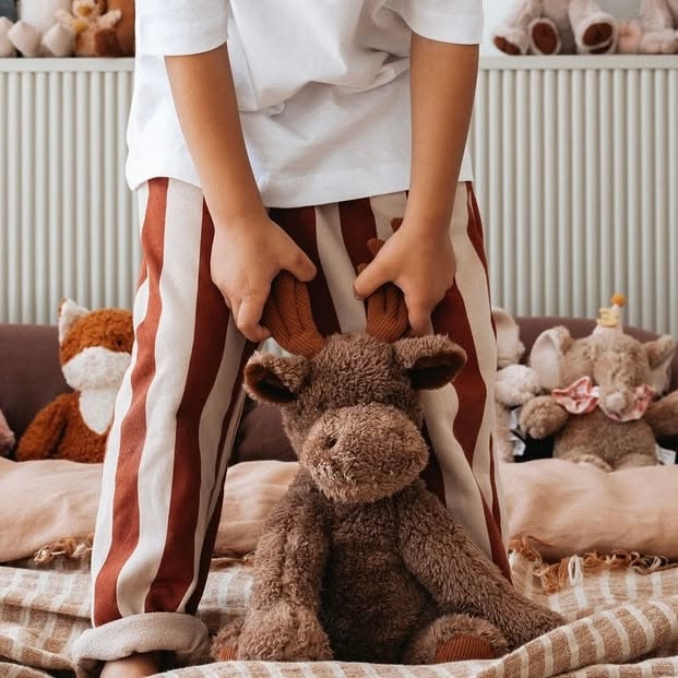 A child wearing a white t-shirt and rust-and-cream vertically striped pajama pants sits on a cozy bed surrounded by plush toys, holding a brown plush moose toy.