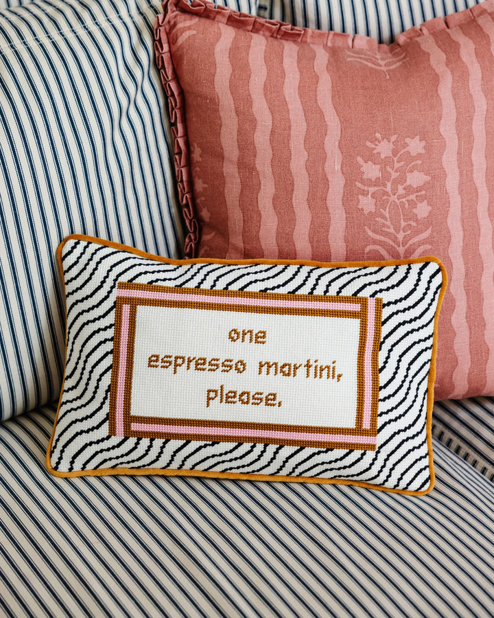 A rectangular decorative throw pillow with a black and white zebra print border, a rust-orange inner border, and embroidered brown text reading 'one espresso martini, please.' It is styled on a striped sofa with pink and blue pillows in the background.