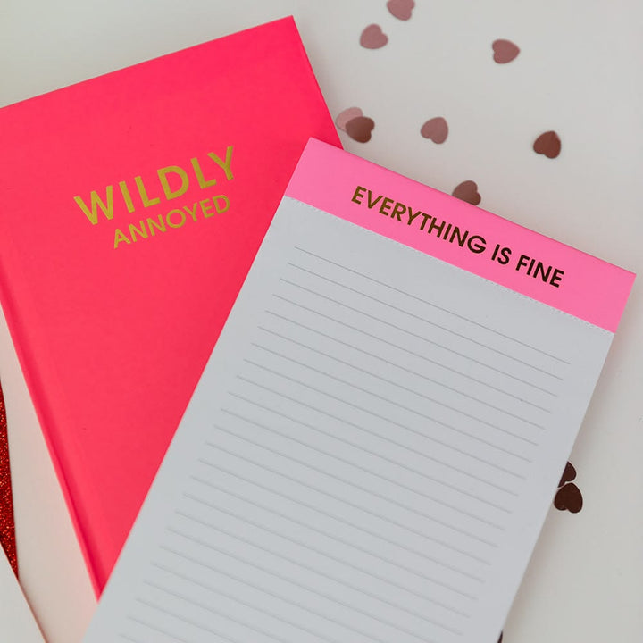 Notebook with 'EVERYTHING IS FINE' on pink cover and 'WILDLY ANNOYED' on red cover.
