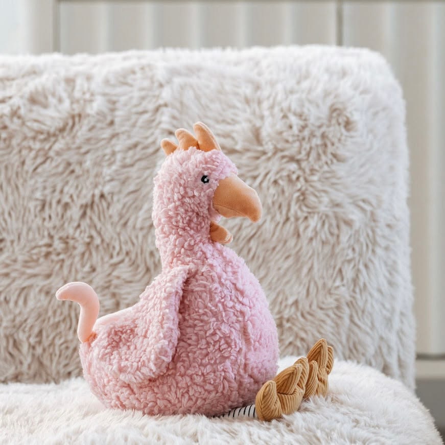A pink textured plush chicken toy with a peach beak and feet, sitting on a white faux fur surface in front of a fluffy cream pillow.