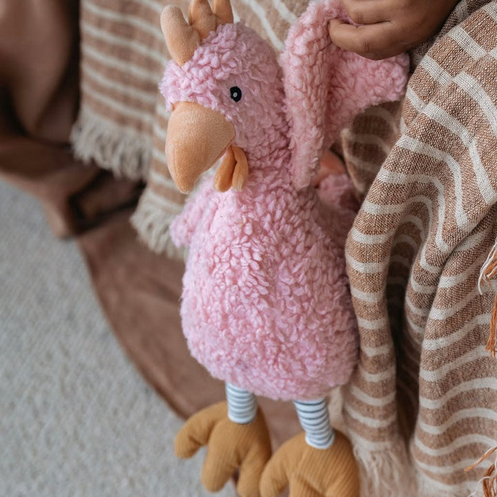 A pink fluffy stuffed bird toy with a peach beak, striped legs, and tan feet, being held by a person wrapped in a brown and cream striped blanket.