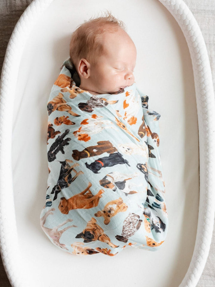A sleeping newborn baby swaddled in a light blue muslin swaddle blanket with a colorful print of various dog breeds, resting inside a white oval bassinet.