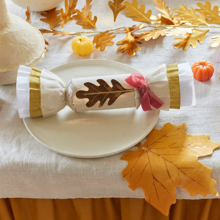 A white paper party cracker with a brown oak leaf design in the center, wrapped with white and gold striped bands and tied with a pink ribbon, displayed on a white plate on a tablecloth with autumn decorations.
