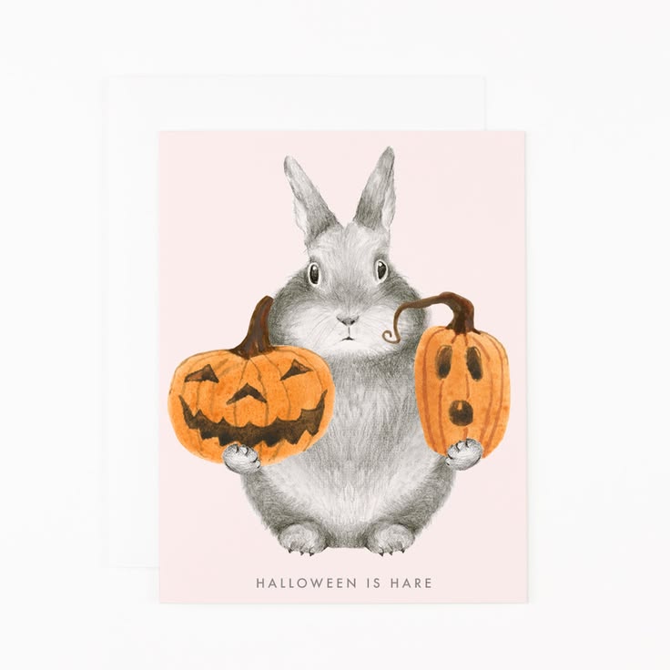 A square greeting card featuring a watercolor-style gray rabbit holding two orange jack-o'-lantern pumpkins, with the text 'HALLOWEEN IS HARE' at the bottom. A white envelope is partially visible behind the card on a white background.