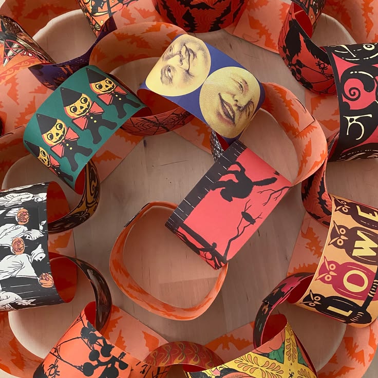 A collection of colorful Halloween-themed paper chains arranged in a circle on a light wooden surface. The chains feature various spooky patterns including jack-o'-lanterns, cats, faces, bats, and the word 'HOMIE' on orange, black, pink, teal, and yellow backgrounds.
