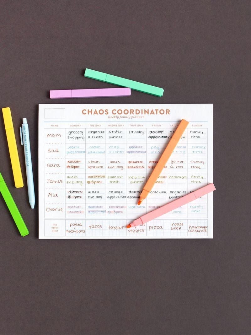 A flat-lay view of a 'Chaos Coordinator' tracking sheet on a dark brown background, surrounded by colorful highlighters.