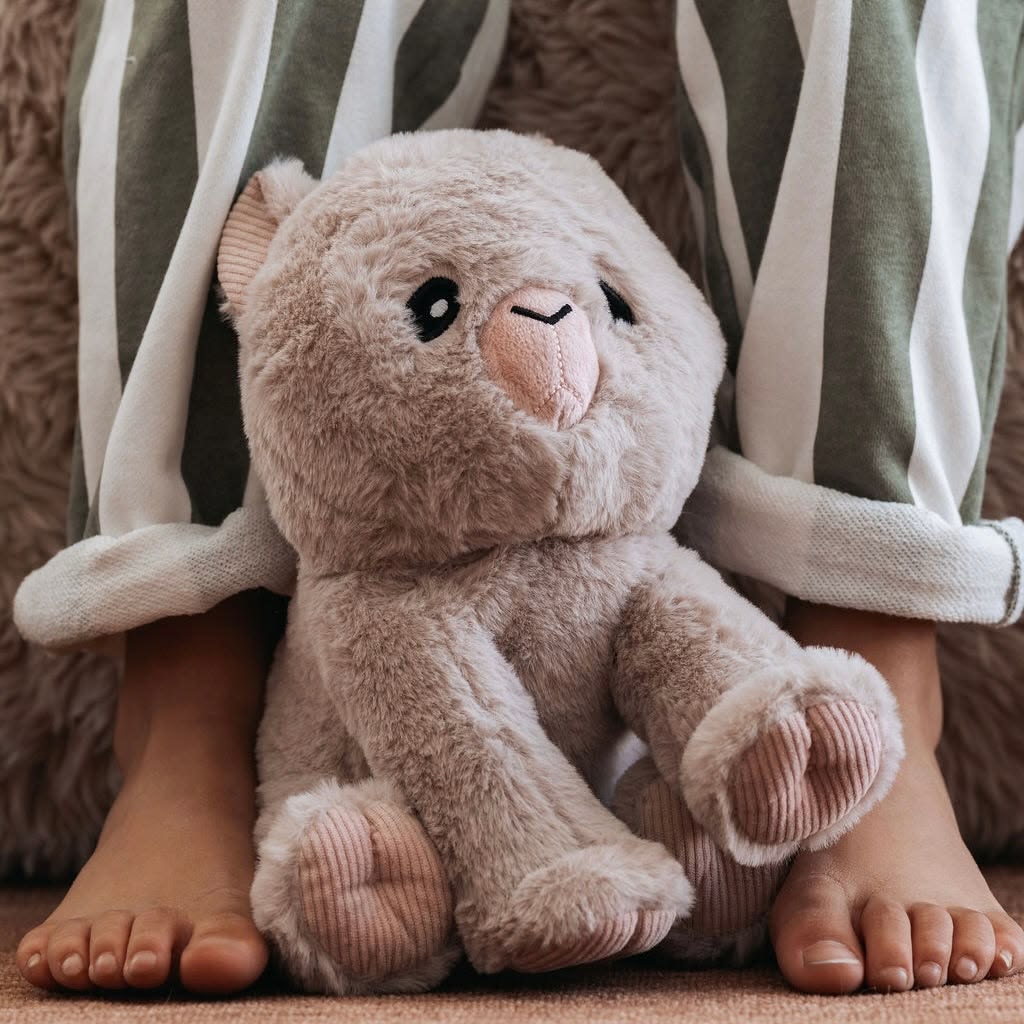 A plush teddy bear toy in a warm taupe-beige color sits on the lap of a person wearing green and white striped pajama pants. The toy features a soft, fuzzy texture, a pink stitched nose and mouth, and corduroy-style paw pads.