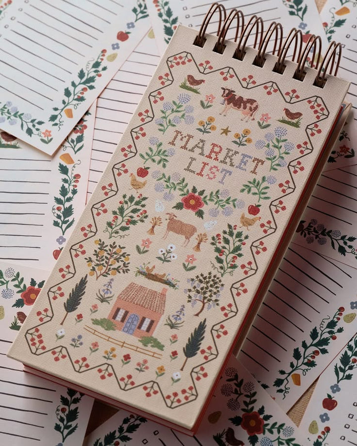 A spiral-bound market list notepad with a whimsical embroidered-style cover illustration, placed on top of matching floral lined paper sheets.